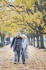 Ласковая пожилая пара, гуляющая среди деревьев и листьев в осеннем парке — стоковое фото