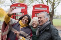 Senior friends taking selfie in front of red telephone booths in autumn park — Stock Photo