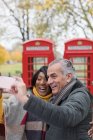 Feliz pareja de ancianos tomando selfie frente a cabinas telefónicas rojas en el parque de otoño - foto de stock