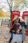 Senior women friends with shopping bags taking selfie in autumn park in front of red telephone booths — Stock Photo