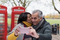 Casal sênior beijando e tirando selfie no parque na frente de cabines telefônicas vermelhas — Fotografia de Stock