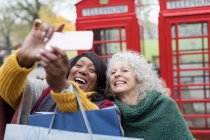 Smiling senior women friends taking selfie in park in front of red telephone booths — Stock Photo