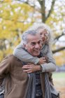 Playful, smiling senior couple piggybacking in autumn park — Stock Photo