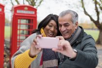 Sorrindo casal sênior tomando selfie no parque na frente da cabine telefônica vermelha — Fotografia de Stock