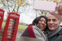 Affectionate senior couple taking selfie in autumn park in front of red telephone book — Stock Photo