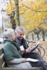 Старшая пара читает газету и пьет кофе на скамейке запасных в осеннем парке — стоковое фото
