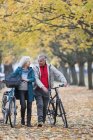 Sorrindo casal sênior caminhando bicicletas entre árvores e folhas no parque de outono — Fotografia de Stock