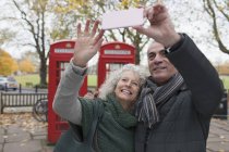 Улыбающаяся пожилая пара делает селфи в осеннем парке перед красными телефонными будками — стоковое фото