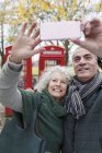 Smiling senior couple taking selfie in park in front of red telephone booths — Stock Photo