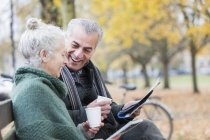Sorridente coppia anziana lettura giornale e bere caffè sulla panchina nel parco autunnale — Foto stock