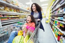 Играющая мать толкает смеющуюся дочь в корзине в супермаркете — стоковое фото