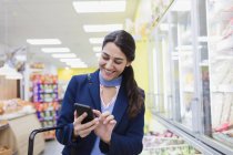 Mujer sonriente con compras de teléfonos inteligentes en el supermercado - foto de stock