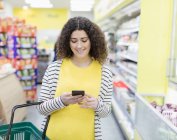 Mujer sonriente con compras de teléfonos inteligentes en el supermercado - foto de stock