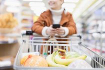 Donna con smart phone spingendo carrello della spesa nel supermercato — Foto stock