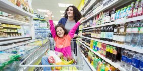 Mother pushing playful daughter in shopping cart in supermarket aisle — Stock Photo