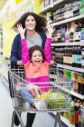Mother pushing excited daughter in shopping cart in supermarket aisle — Stock Photo