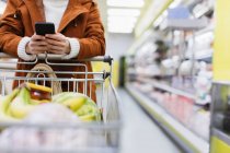 Mujer con teléfono inteligente empujando carrito de compras en el supermercado - foto de stock