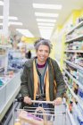 Retrato sonriente, mujer segura empujando carrito de la compra en el pasillo del supermercado - foto de stock