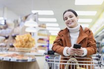 Retrato sorridente mulher com telefone inteligente compras no supermercado — Fotografia de Stock