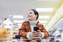 Femme souriante et confiante avec des achats de téléphone intelligent dans un supermarché — Photo de stock