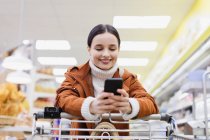 Mulher com telefone inteligente empurrando carrinho de compras no supermercado — Fotografia de Stock