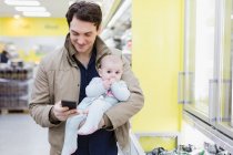 Vater mit kleiner Tochter checkt Smartphone, kauft im Supermarkt ein — Stockfoto