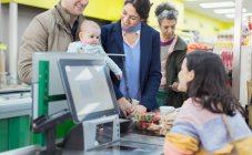 Friendly cashier talking with and helping couple with baby at supermarket checkout — Stock Photo