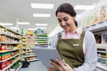 Mulher sorrindo merceeiro usando tablet digital no corredor do supermercado — Fotografia de Stock