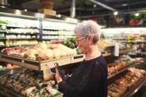 Mujer mayor con compras de teléfonos inteligentes en la sección de productos de supermercado - foto de stock