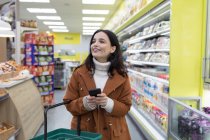 Smiling young woman with smart phone grocery shopping in supermarket — Stock Photo