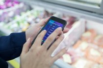 Cerca de la mujer que usa la aplicación de lista de compras de teléfonos inteligentes en el supermercado - foto de stock