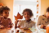 Portrait happy boy with autumn leaf doing crafts with sisters at table — Stock Photo