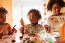 Smiling boy with autumn leaf doing crafts with sisters at table — Stock Photo
