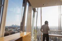 Empresário falando no telefone inteligente na janela do arranha-céus moderno ensolarado — Fotografia de Stock
