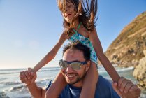 Счастливый отец несет дочь на плечах на солнечном пляже — стоковое фото