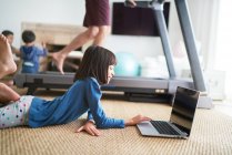 Chica usando el ordenador portátil en el suelo junto a padre en la cinta de correr - foto de stock