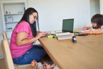 Bambini che giocano e fanno i compiti mentre la madre lavora a tavola — Foto stock