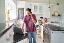 Man working at laptop in kitchen with kids eating — Stock Photo