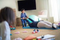 Ragazzo che gioca con la madre che si esercita sul tappetino yoga in soggiorno — Foto stock
