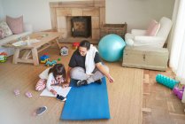 Мама на коврике для йоги смотрит, как дочь раскрашивает пол в гостиной — стоковое фото