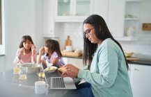 Madre trabajando en el ordenador portátil en la cocina mientras las hijas desayunan - foto de stock