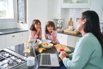 Hijas felices desayunando y viendo a la madre trabajar en la cocina - foto de stock