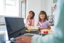 Glückliche Schwestern frühstücken in der Küche, während die Mutter am Laptop arbeitet — Stockfoto
