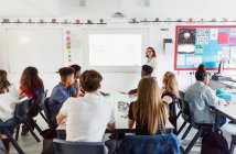 Estudiantes de secundaria observando a una maestra dirigiendo la lección en la pantalla de proyección en el aula - foto de stock