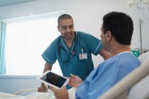 Чоловіча медсестра розмовляє з пацієнтом, використовуючи цифровий планшет у лікарняному ліжку — стокове фото