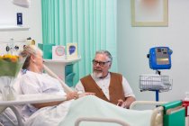 Ласковый пожилой мужчина навещает, разговаривает с женой, отдыхающей в палате больницы — стоковое фото