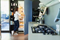 Жінка стоїть у відкритому холодильнику на кухні — стокове фото