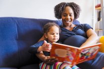 Schwangere Mutter liest Tochter Buch auf Sofa vor — Stockfoto