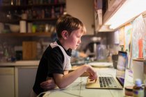 Мальчик делает домашнее задание за ноутбуком на кухне — стоковое фото
