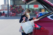 Donna con maschera viso carico generi alimentari in auto nel parcheggio — Foto stock